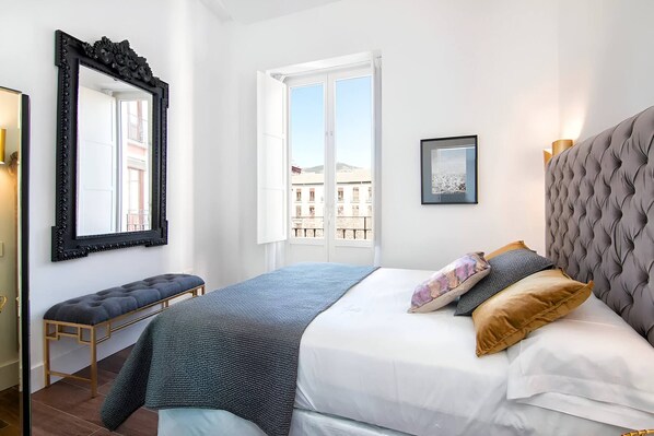 A bedroom with a large bed, a bench, a window with a view, and a framed picture on the wall.