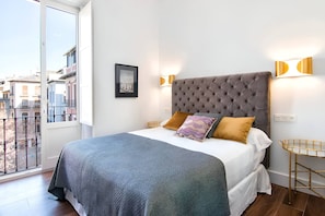 Une chambre avec un grand lit, une fenêtre donnant sur des immeubles et une petite table ronde avec une lampe.