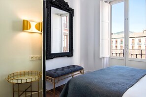 A bedroom with a bed, a bench, a mirror, and a window with a view of buildings.