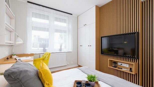 A modern bedroom with a bed, a TV mounted on a wooden panel wall, a window with sheer curtains, and a bedside table with a lamp and a tray with cups and a plant. - Spacious Apartment | 2 Bedrooms | In the Heart of Kazimierz. (Kraków)
