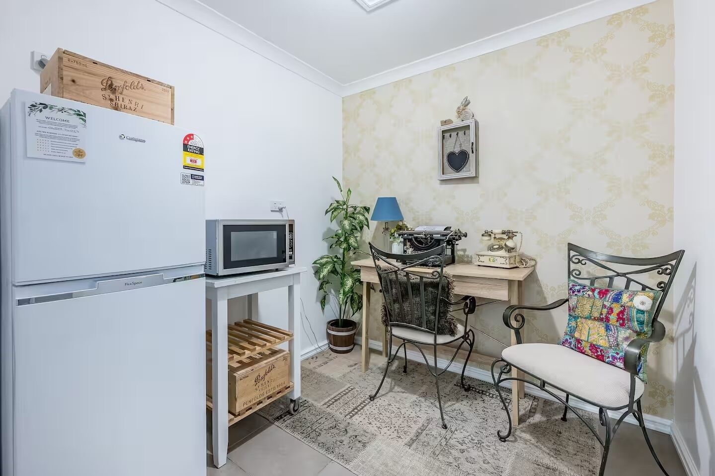 Ett kompakt kök med ett vitt kylskåp, en mikrovågsugn, ett litet träbord, en gammal skrivmaskin och en metallstol med en färgglad kudde.