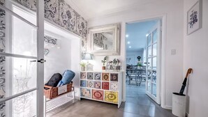 Un couloir avec un papier peint à motifs, un panier en osier contenant divers objets, un meuble décoratif orné de bibelots, et une porte donnant sur une autre pièce.