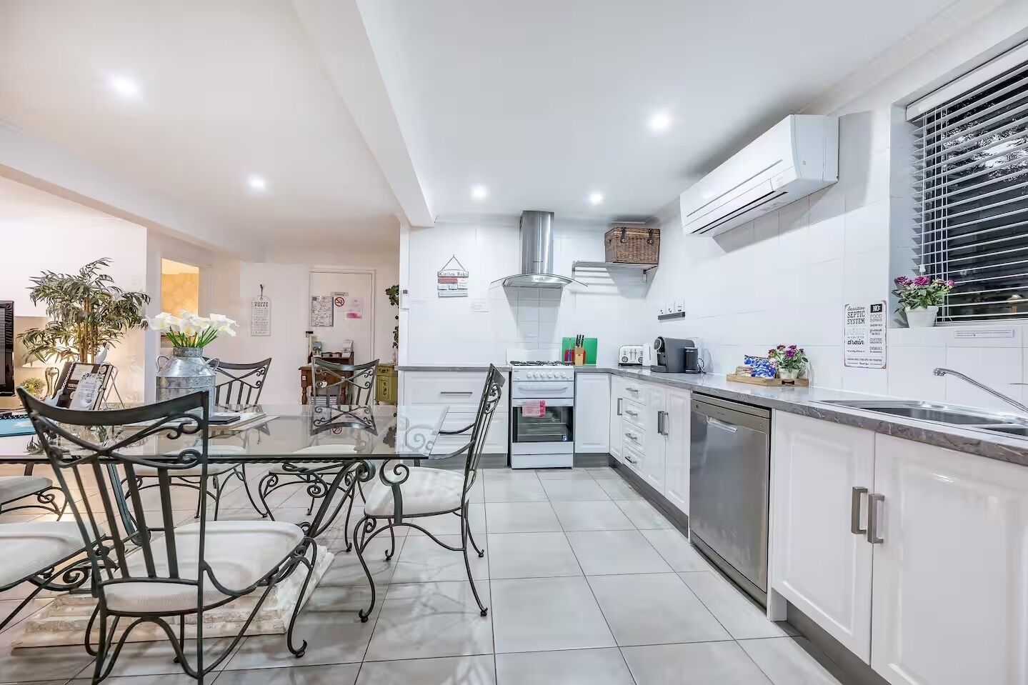 Una cucina moderna con mobili bianchi, un lavello in acciaio inossidabile e un piano cottura a gas. C'è una zona pranzo con un tavolo in vetro e sedie metalliche.