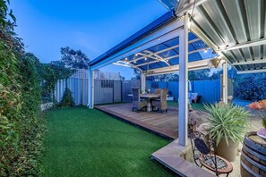 A covered patio with a wooden table and chairs, surrounded by a well-maintained lawn and a metal fence.