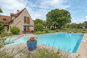 A swimming pool with a stone house, outdoor seating, and a large tree.