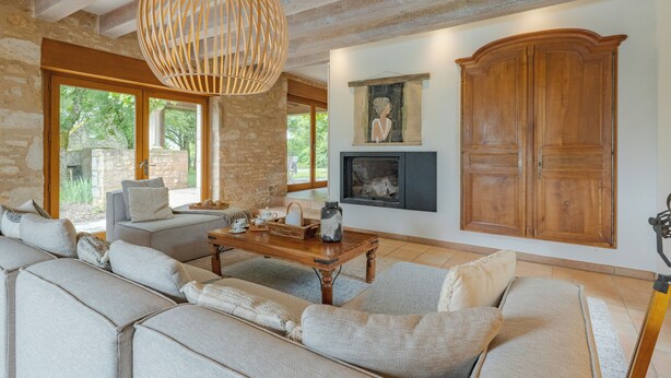 A spacious living room with a large sofa, a wooden coffee table, a hanging light fixture, a fireplace, and a stone wall.