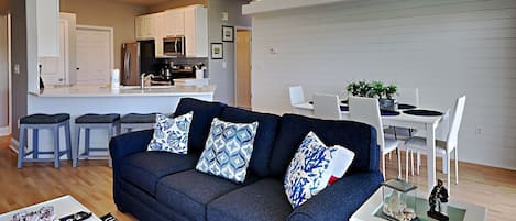 Una sala moderna con un sofá azul oscuro, una mesa de comedor blanca y una cocina al fondo.