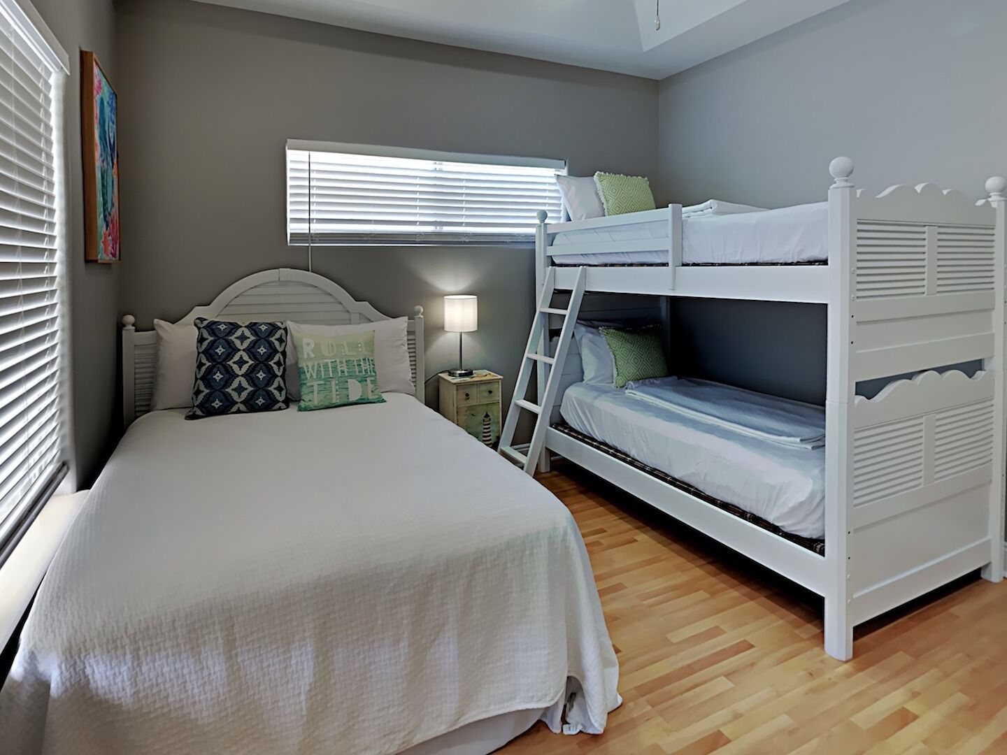 Un dormitorio con una cama individual, una litera, una mesita de noche con lámpara y una ventana con persianas.