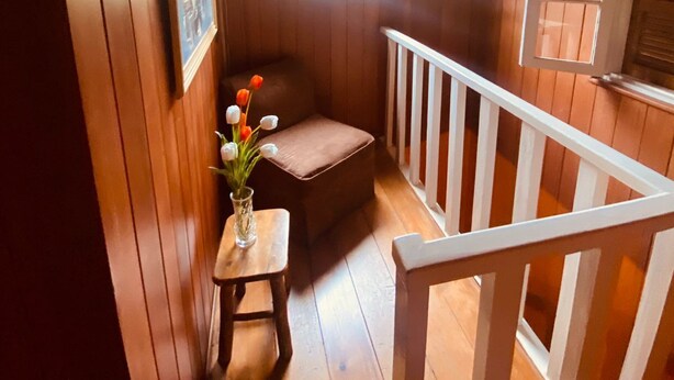 A wooden cabin interior with a small seating area, a window with shutters, and a white railing. - Casa Fontana (Campos do Jordão)