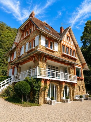 Een groot, twee verdiepingen hoog stenen huis met een balkon, schoorsteen en een kleine tuin.