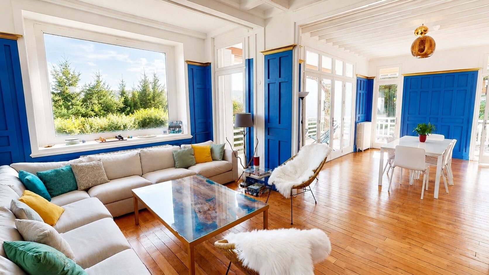 Un salon spacieux avec une grande fenêtre, un canapé, une table basse en verre et un coin repas avec une table et des chaises blanches.