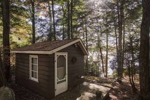Une petite cabane en bois, dotée d’une porte et d’une fenêtre, entourée de grands arbres et agrémentée d’une terrasse.