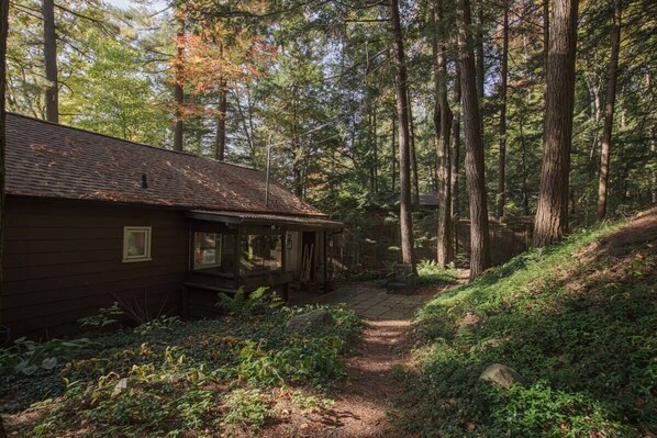 A cabin with a covered porch, surrounded by a forested area with a dirt path leading to it.