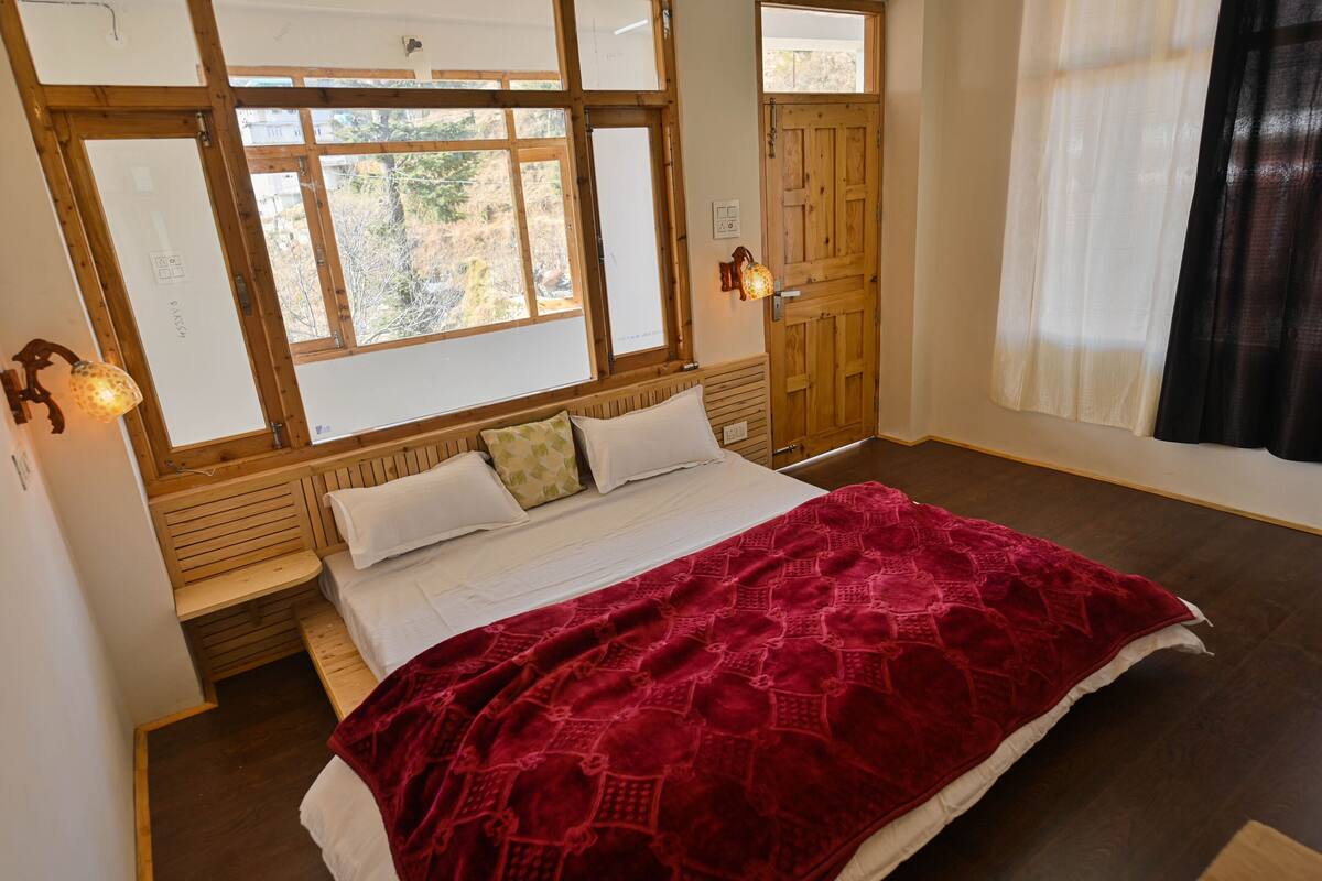 A bedroom with a wooden headboard, a large window with a view of trees, a bed with a red blanket, and a wooden bench at the foot of the bed.