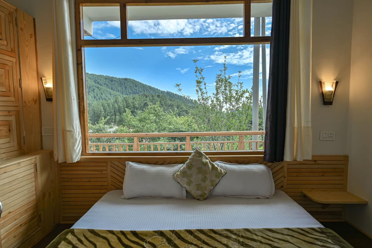 A wooden cabin room with a bed, a window with a view of a forested mountain, and a wooden bench.