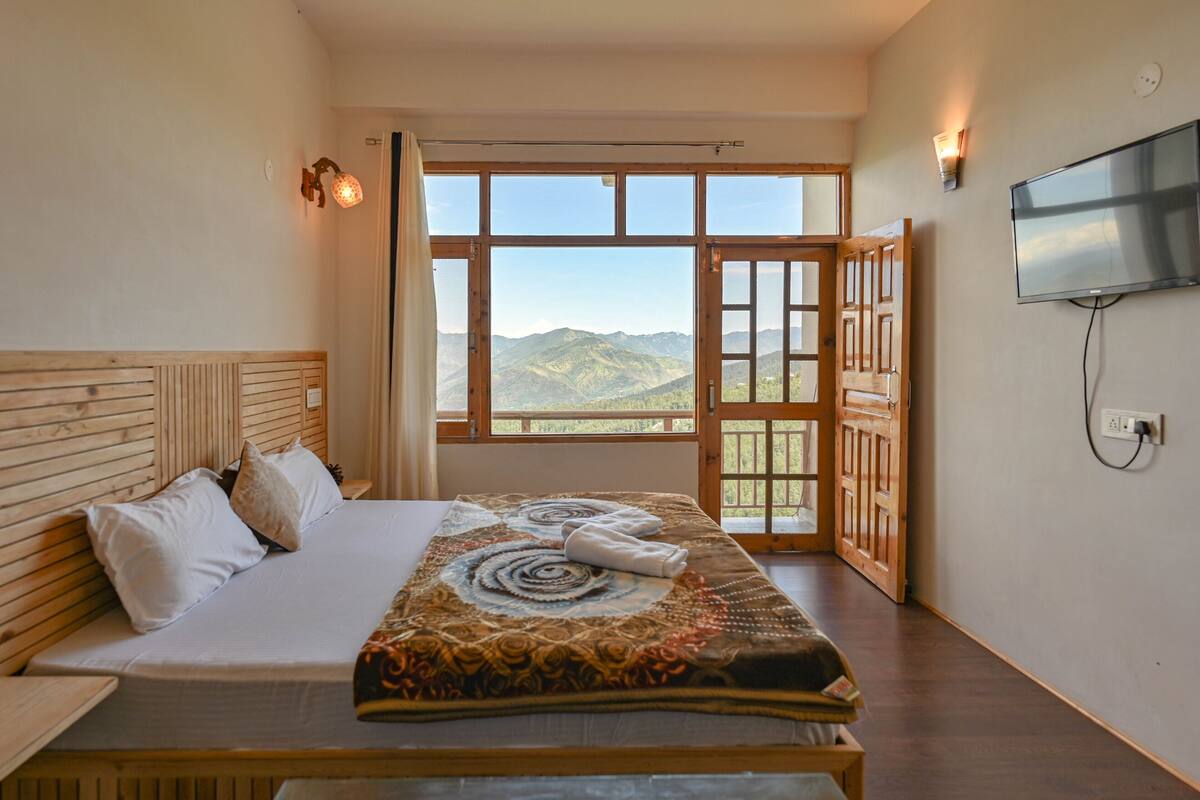 A bedroom with a large bed, a TV mounted on the wall, and a view of mountains through a large window.