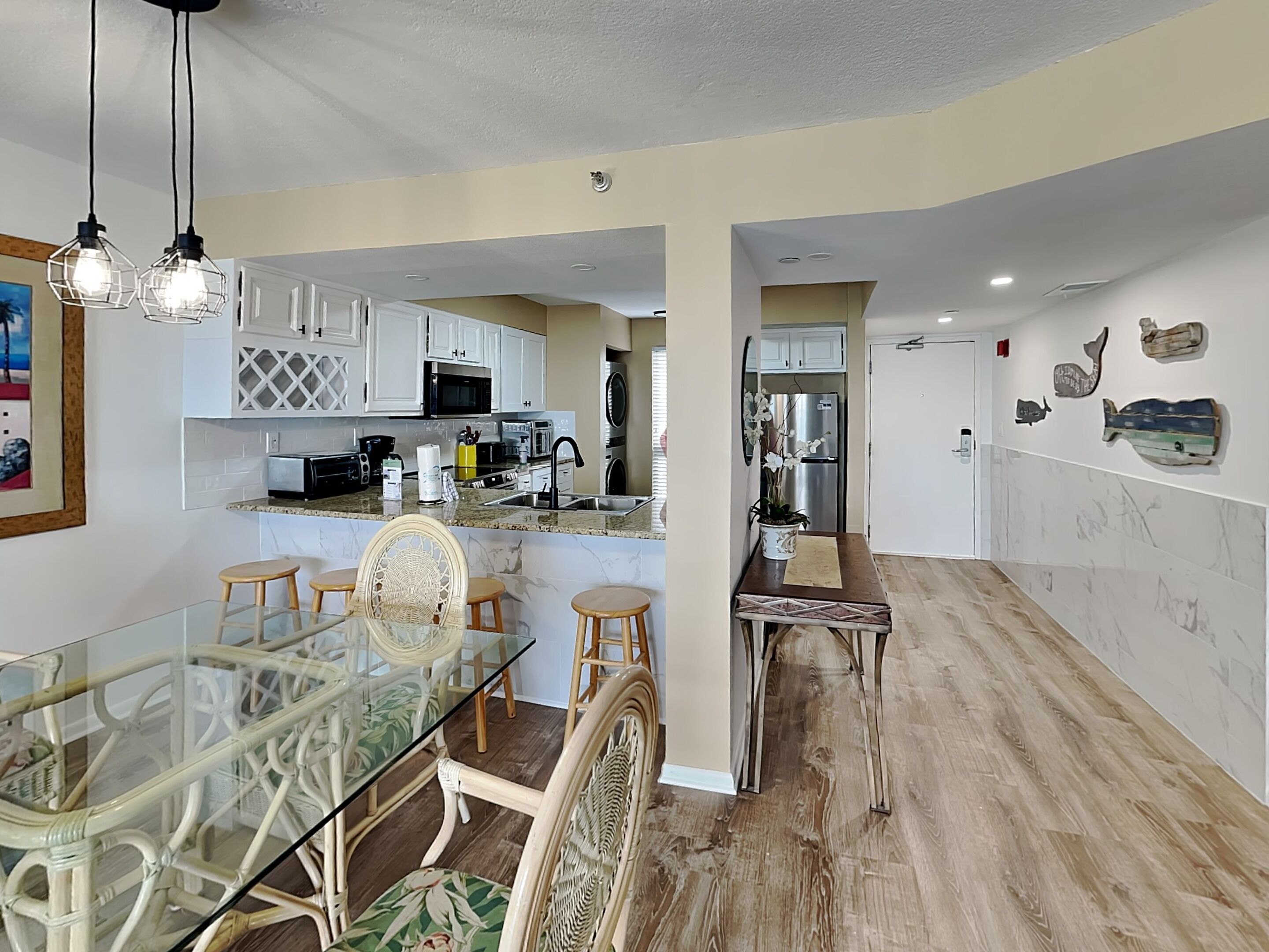 Una cocina moderna con armarios blancos, una zona de comedor con una mesa de vidrio y sillas de mimbre, y piso de madera.