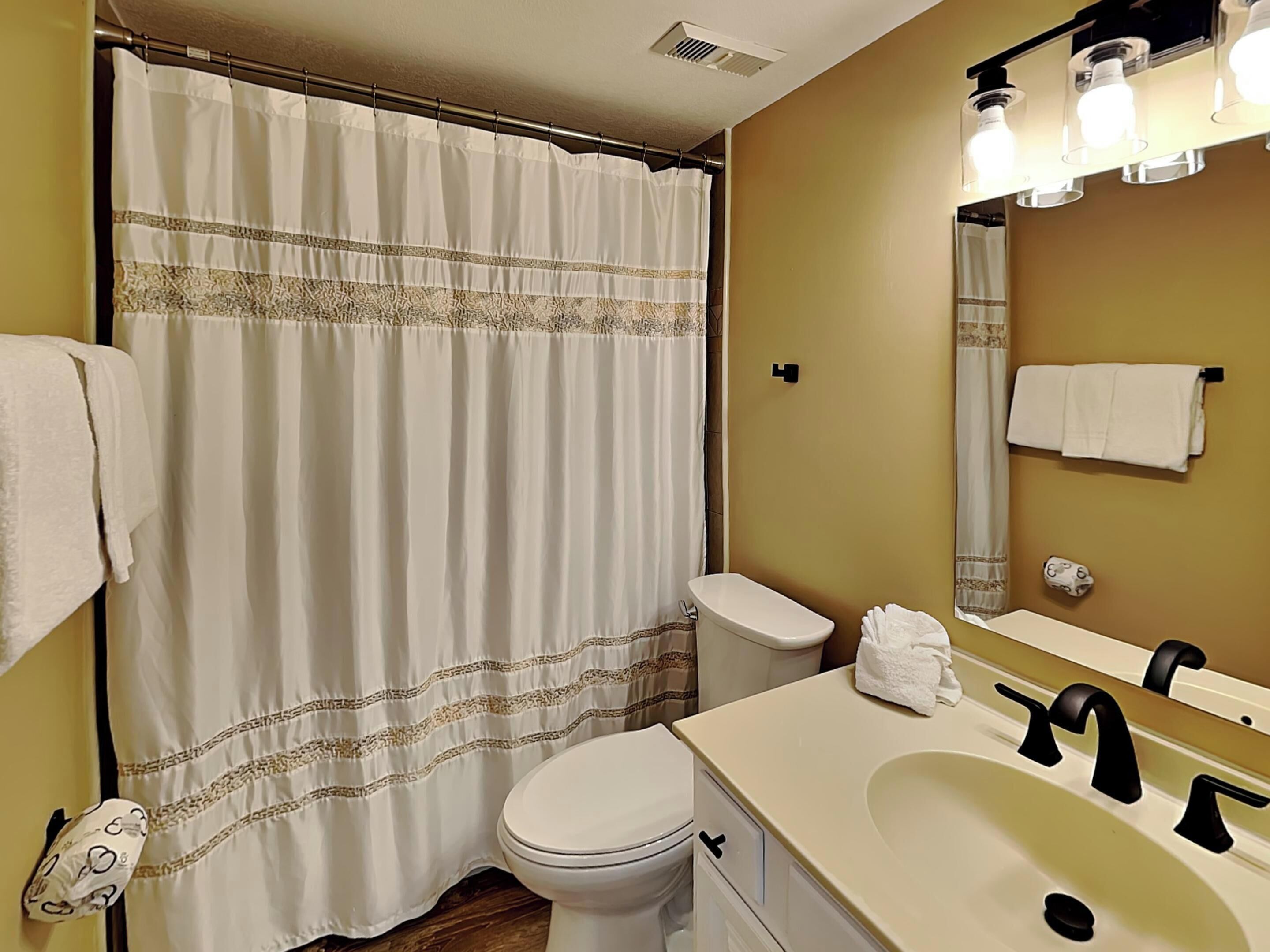 Une salle de bain avec des toilettes blanches, un lavabo surmonté d’un miroir et un rideau de douche orné d’un motif décoratif.