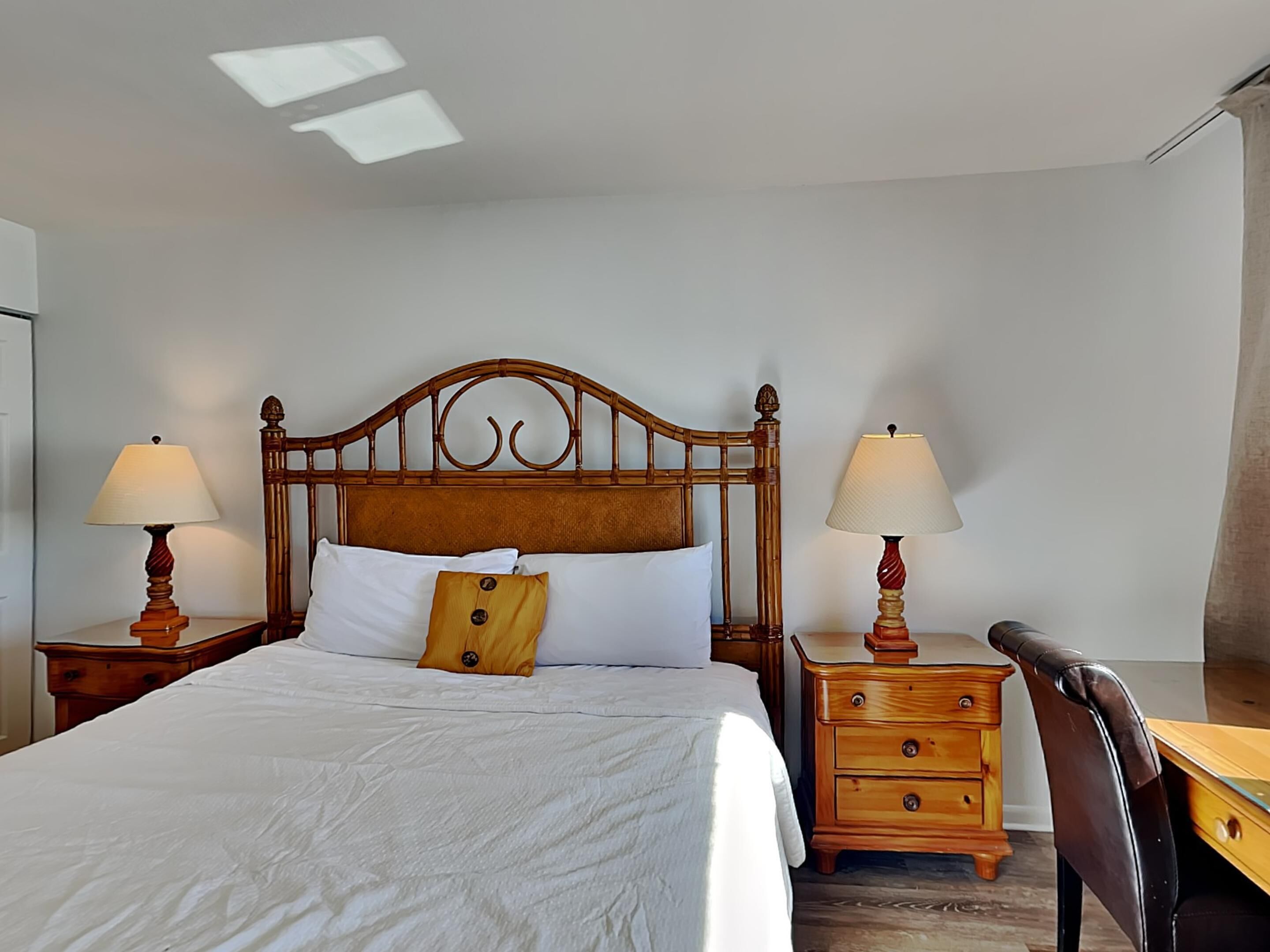 Una cama bien tendida con sábanas blancas, un cabecero de madera, dos mesitas de noche con lámparas, un escritorio de madera y una silla.