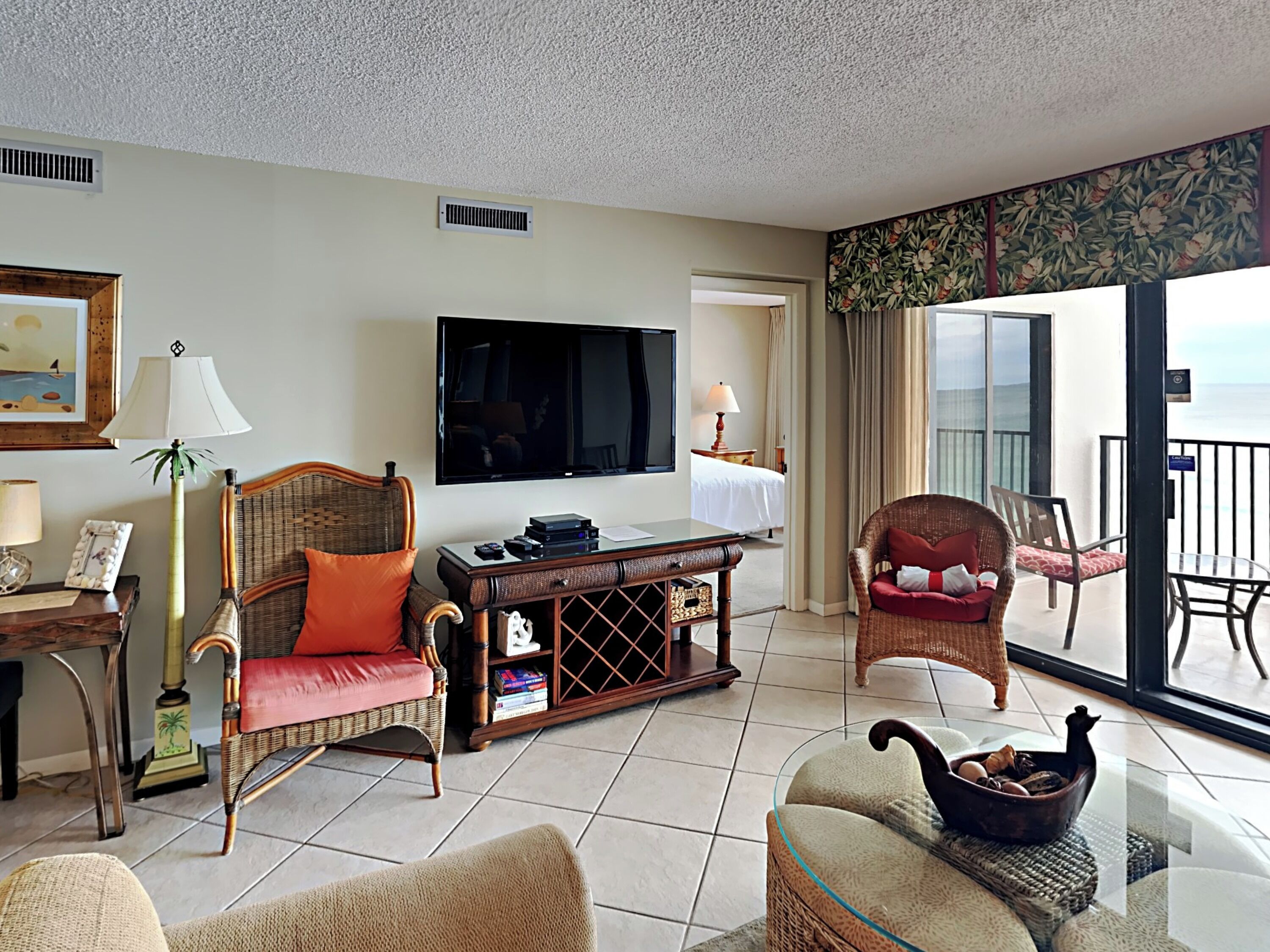 A hotel room with a large flat-screen TV, wicker furniture, a glass coffee table, and a balcony with a view.