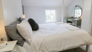 A bedroom with a large bed, a dresser with a mirror, and a window with a view of greenery.