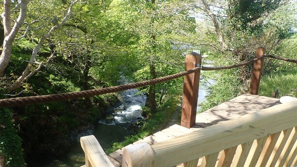 A wooden bridge with a rope railing, leading over a small stream surrounded by greenery. - Beautiful 15th Century Watermill - Lovely Location (Machynlleth)