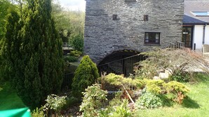 A stone building with a small window and a door, surrounded by lush greenery and a stone archway. - Beautiful 15th Century Watermill - Lovely Location (Machynlleth)