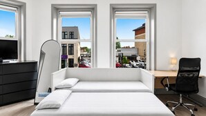 Un dormitorio con dos camas, un escritorio, una silla, un televisor y ventanas amplias con vistas a edificios y coches aparcados.