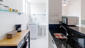 A modern kitchen with a coffee maker, washing machine, sink, and microwave. - BR06 Quarto e Sala - 550m Praia da Barra (Salvador)