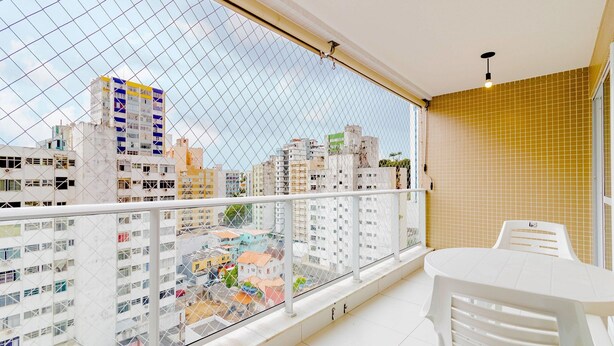 A balcony with a view of a cityscape, a white table, and a white chair. - BR06 Quarto e Sala - 550m Praia da Barra (Salvador)