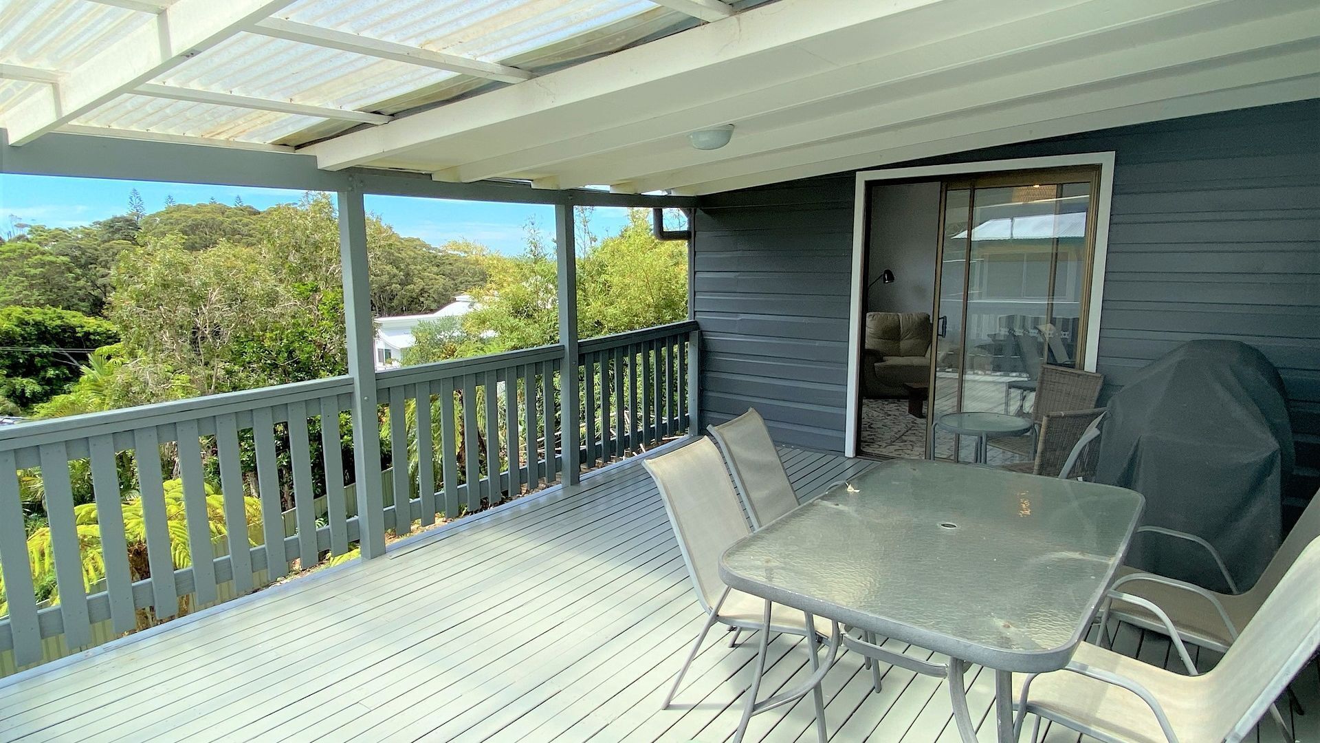 A covered outdoor patio with a table and chairs, a glass door leading to an interior space, and a view of greenery and buildings in the background.