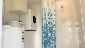 A compact bathroom with a washing machine, a shower with a handheld showerhead, and a curtain with a leaf pattern. - Bluebell - Walking distance to the beach, suitable for large families, and within minutes drive to a (Nambucca Heads)