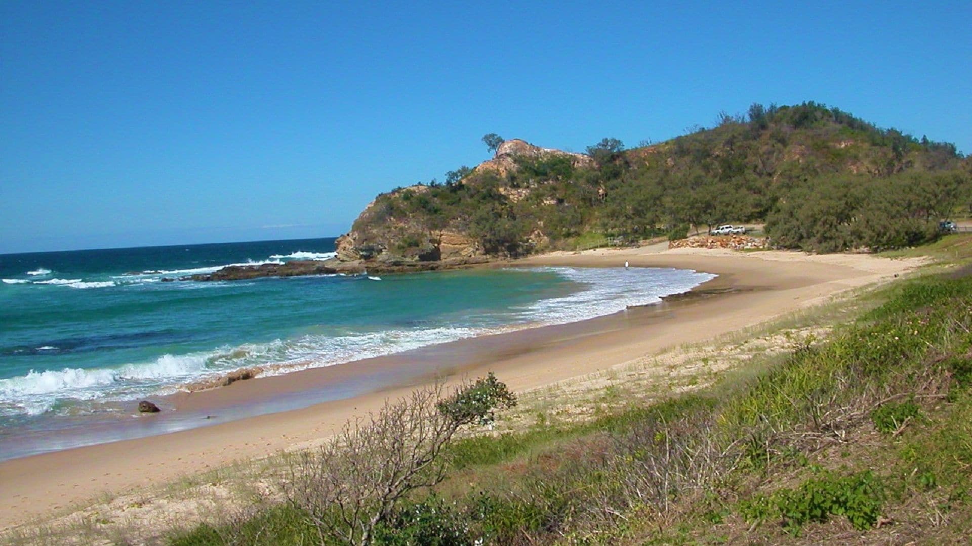 A coastal landscape with a sandy beach, a rocky outcrop, and a hill with vegetation.