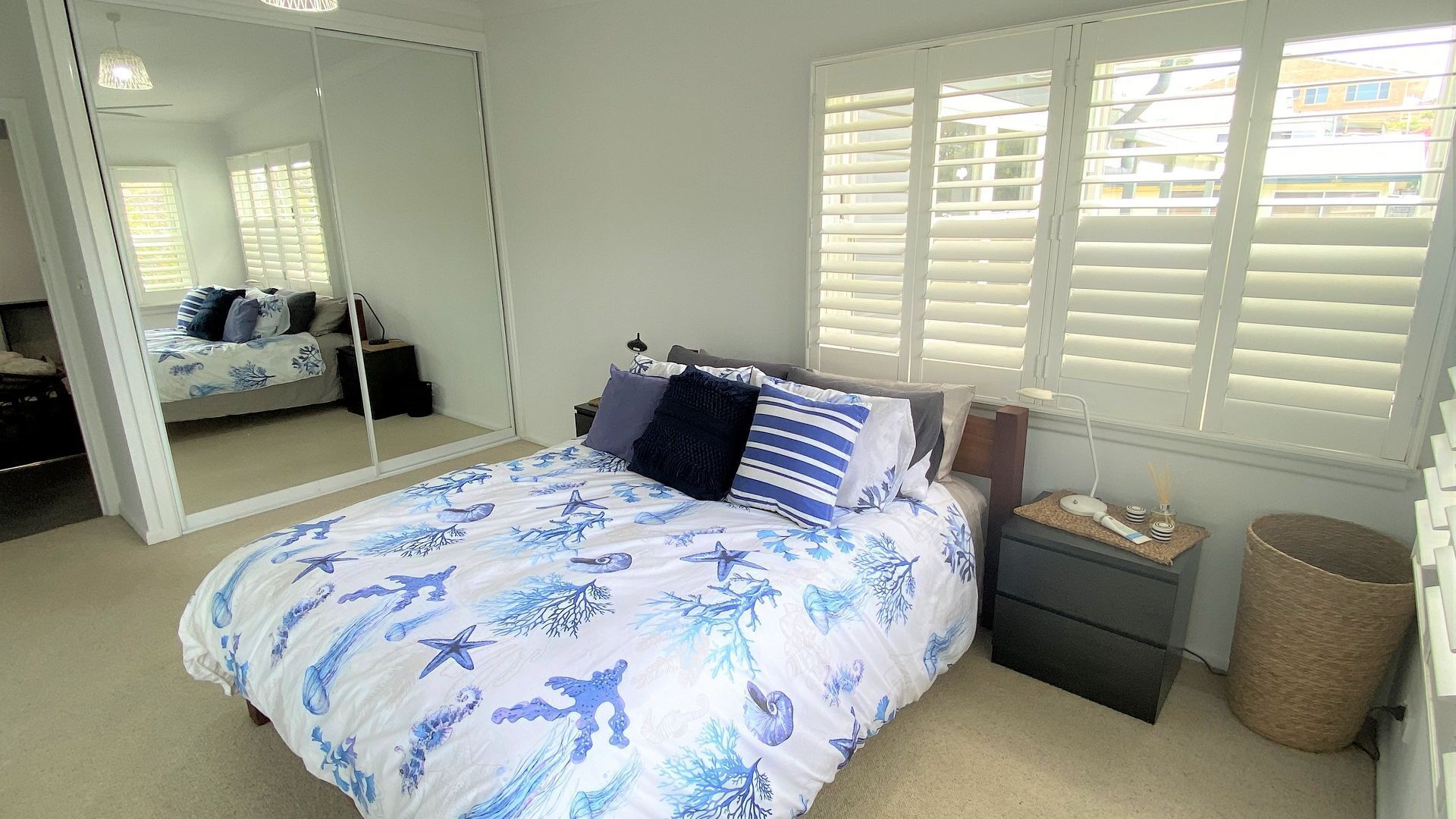 A bedroom with a bed, a bedside table, a mirror, and a window with blinds.