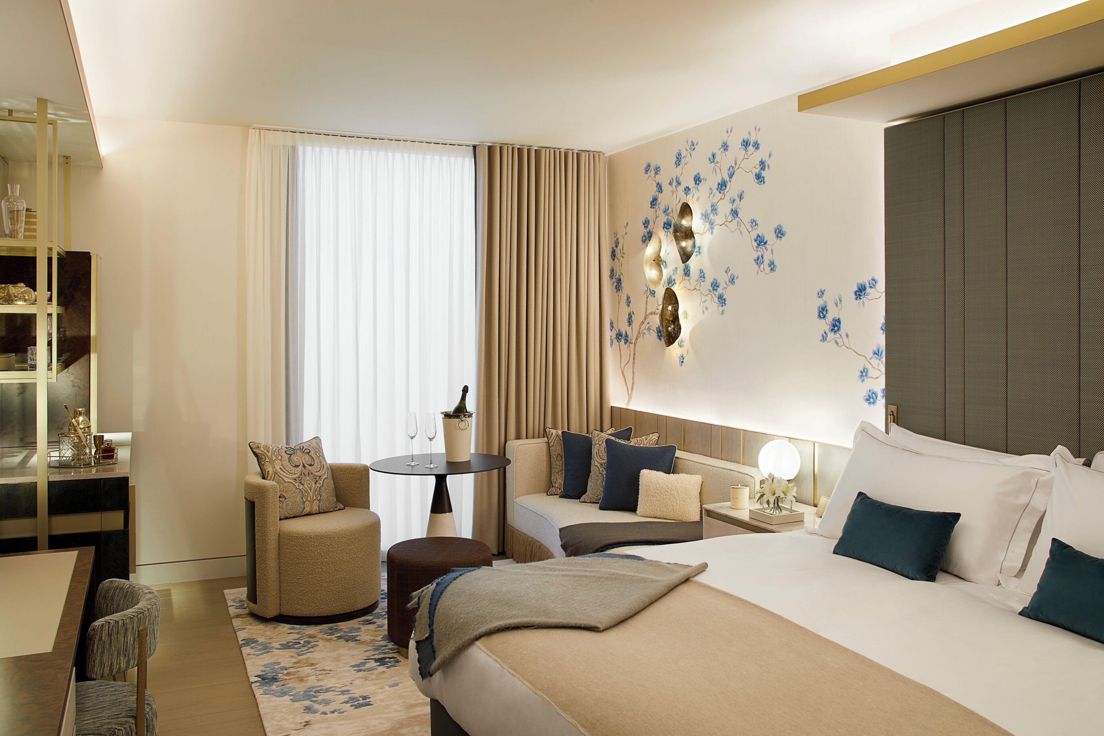A hotel room with a bed, two armchairs, a small table, a bookshelf, and a large window with curtains.