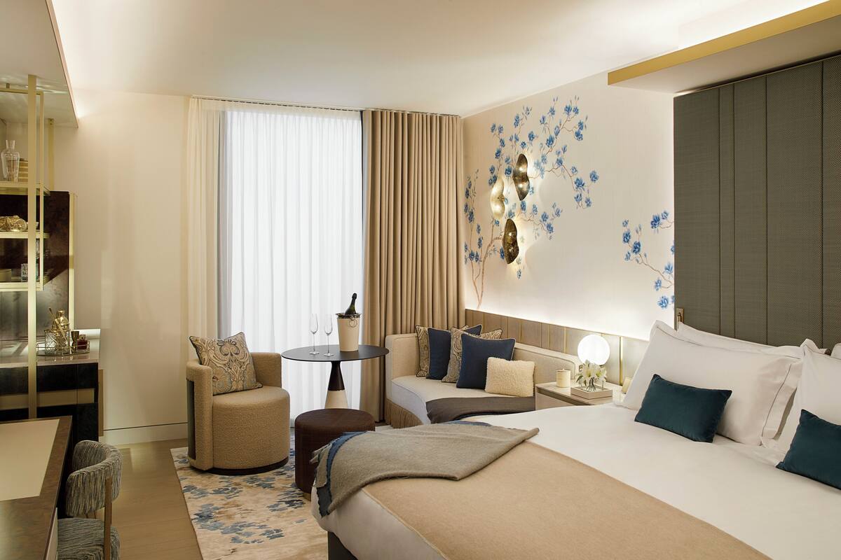 Une chambre d’hôtel comprenant un lit, deux fauteuils, une petite table, une bibliothèque et une grande fenêtre avec des rideaux.