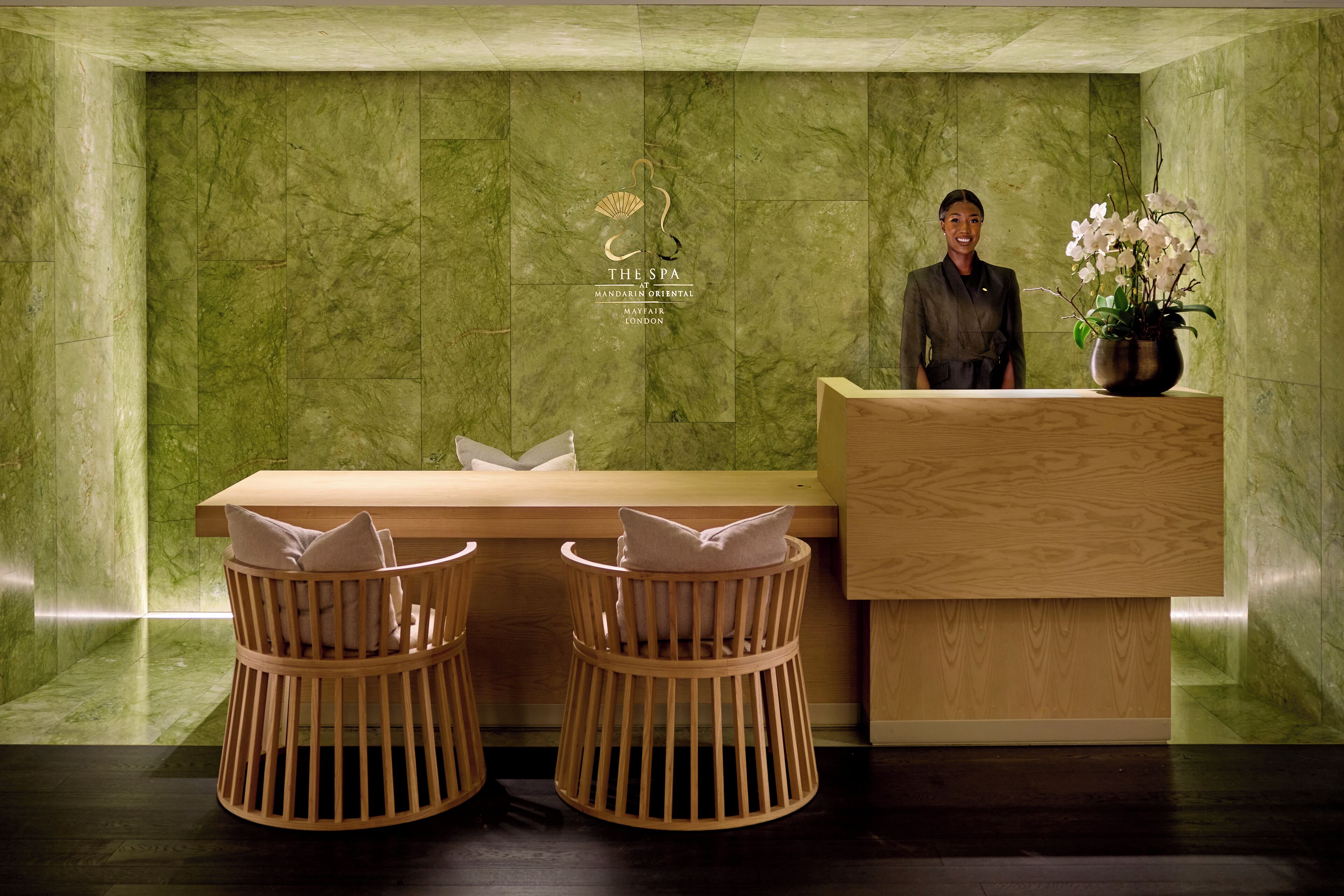 A reception area with a wooden desk, two wicker chairs, a potted orchid, and a wall with a logo.