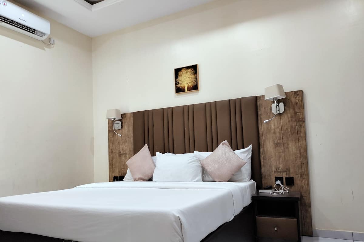 A hotel room with a large bed, a wooden headboard, bedside lamps, a nightstand, and a wall-mounted air conditioner.