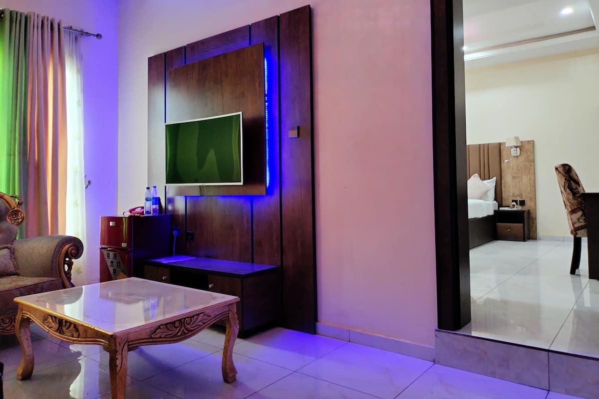 A hotel room with a flat-screen TV mounted on a wooden panel, a glass coffee table, a sofa, and a bed with a nightstand.
