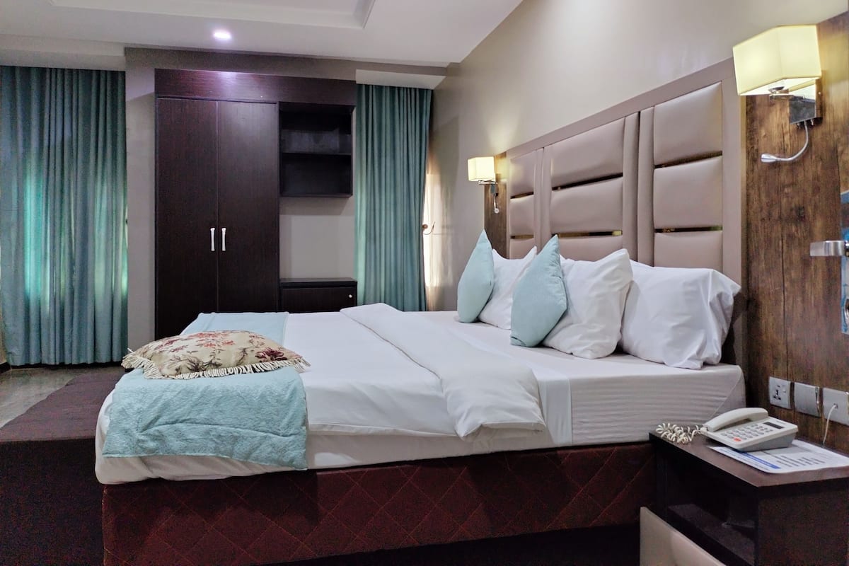 A hotel room with a large bed, a wardrobe, a bedside table with a phone, and a wall-mounted lamp.