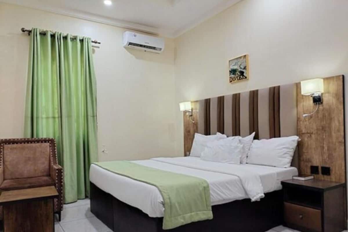 A bedroom with a bed, a chair, a bedside table, a nightstand, an air conditioner, and a wall-mounted lamp.