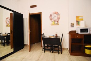 Een kamer met een eettafel, stoelen, een magnetron en een klok aan de muur.