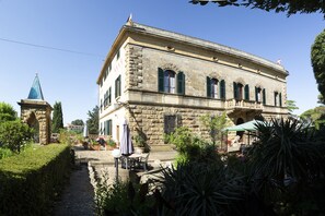 Um edifício de pedra com varanda, uma torre pequena e um jardim com mesa e cadeiras.