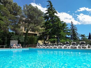 Uma piscina azul límpida com espreguiçadeiras e guarda-sóis, cercada por árvores e um edifício ao fundo.