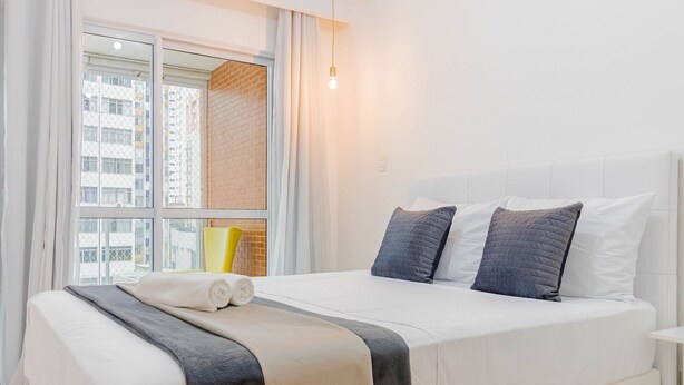 A hotel room with a large bed, white bedding, a window with a view of buildings, and a bedside table. - BR07 Apto a 550m da Praia da Barra (Salvador)