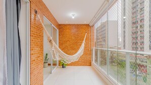 A balcony with a white hammock, a potted plant, and a view of a high-rise building. - BR07 Apto a 550m da Praia da Barra (Salvador)