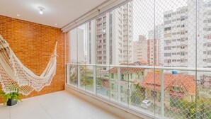 A balcony with a white hammock, a potted plant, and a view of buildings through a glass fence. - BR07 Apto a 550m da Praia da Barra (Salvador)