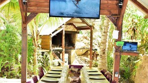 Uma longa mesa de jantar posta para uma refeição em área externa, com vigas de madeira e uma tela de TV acima.
