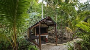 Estrutura rústica ao ar livre com base de pedra e vigas de madeira, cercada por vegetação exuberante e um caminho de pedra.