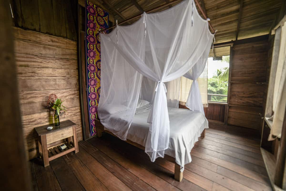 A wooden room with a bed under a white canopy, a small wooden table with a vase, and a colorful wall pattern.